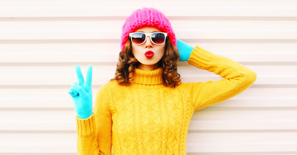 Cooll funny girl blowing red lips wearing colorful knitted yellow sweater pink hat in gloves over white background